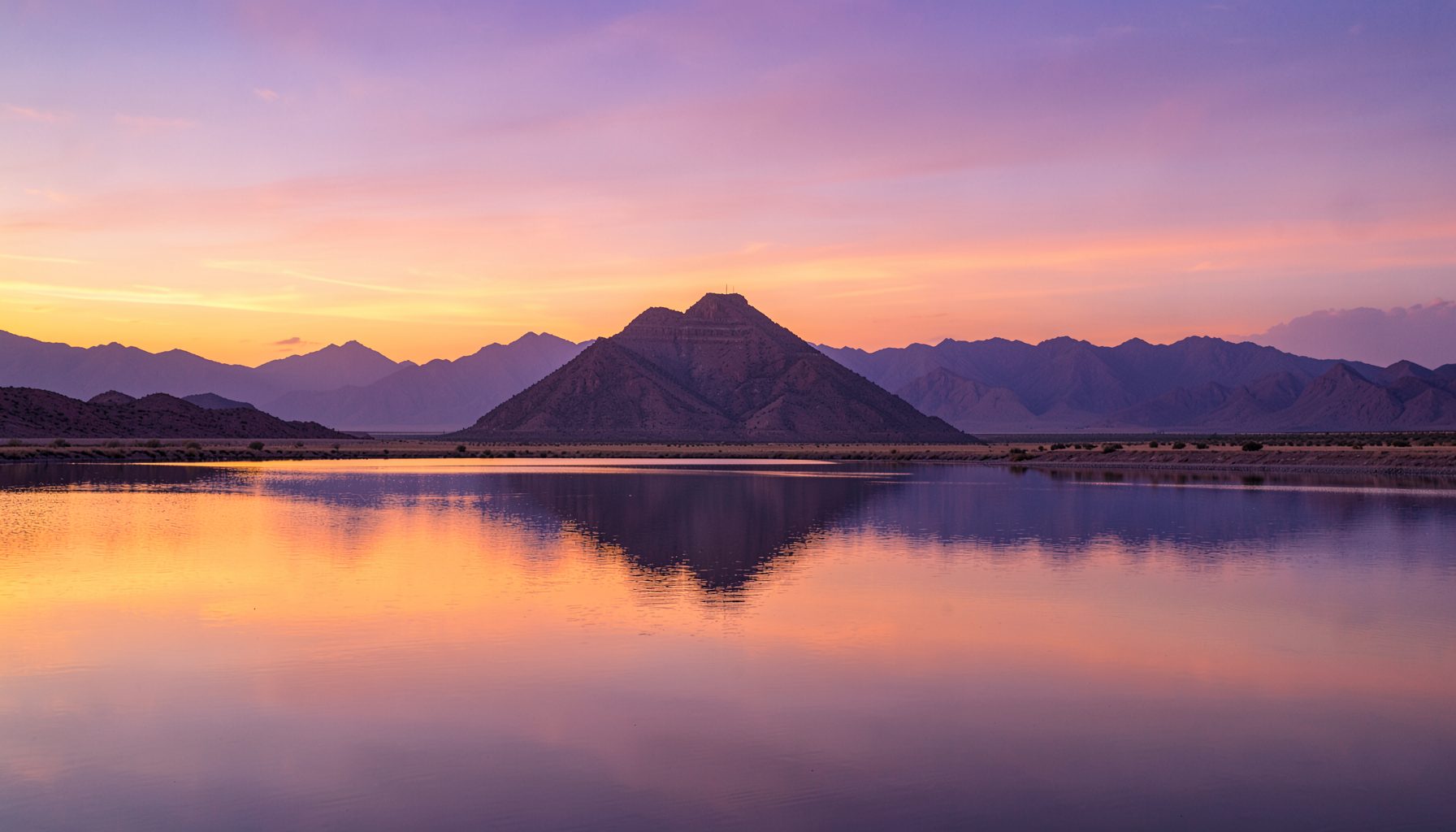 Permukaan Pyramid Lake yang tenang di bawah langit senja berwarna jingga dan ungu, dengan siluet pegunungan gurun di kejauhan